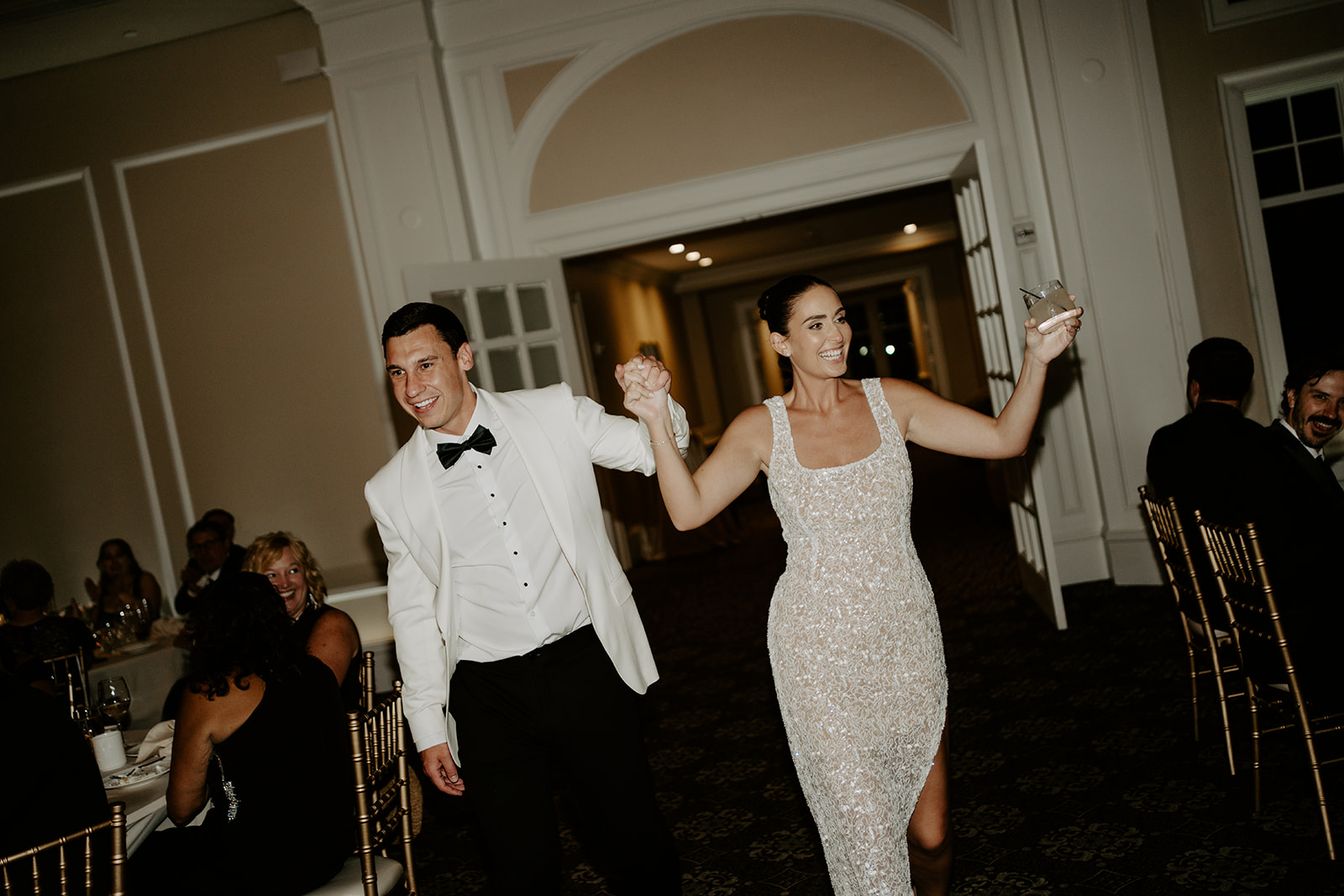 Smiling bride and groom in their reception second outfits entering the hall hand-in-hand.